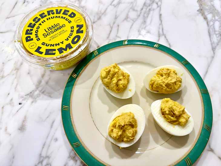 Deviled eggs topped with yellow hummus on a decorative plate next to a container of preserved lemon hummus.