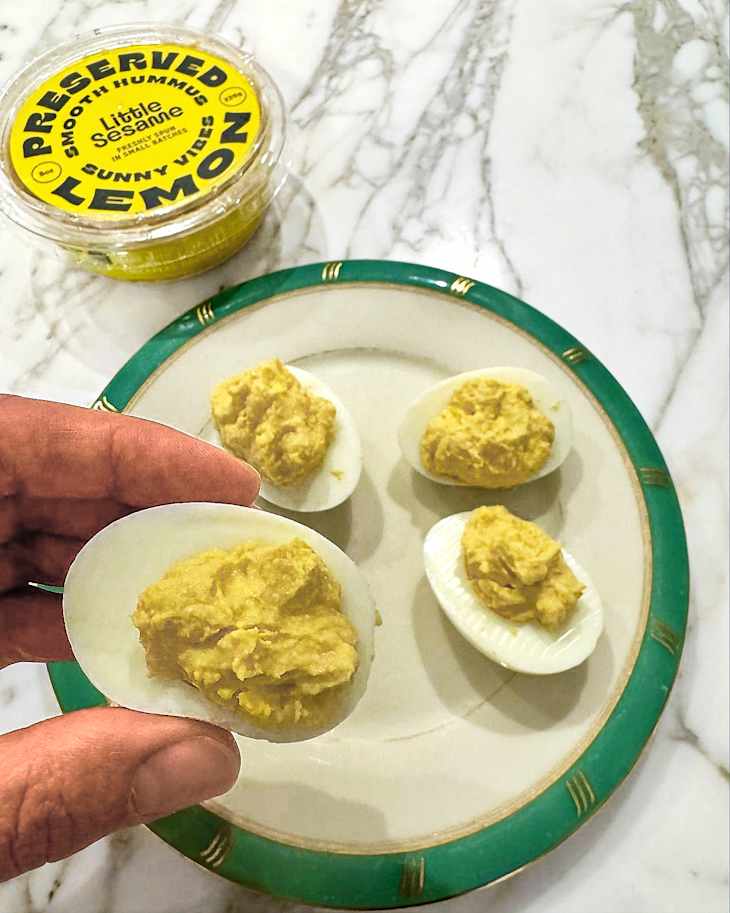 A hand holding a deviled egg topped with hummus, surrounded by more deviled eggs on a decorative plate and a container of preserved lemon hummus.