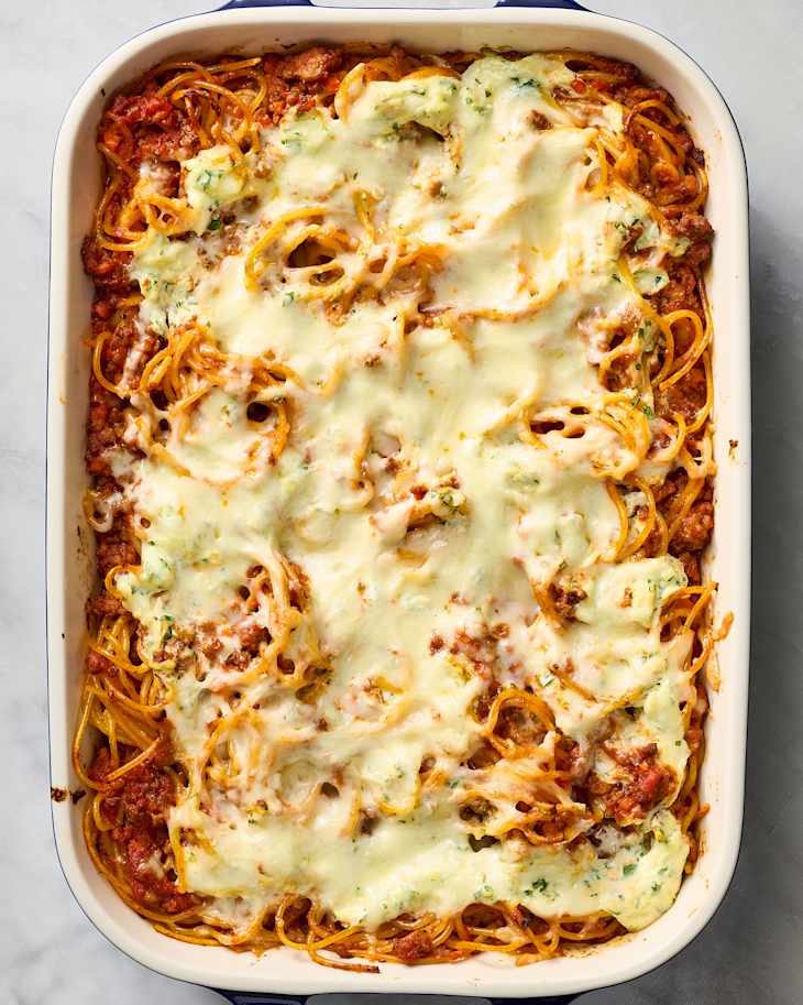 Baked spaghetti casserole topped with melted cheese and herbs in a white dish.