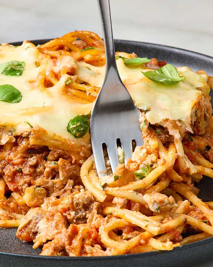 Baked spaghetti with melted cheese, ground meat, and fresh basil on a dark plate.