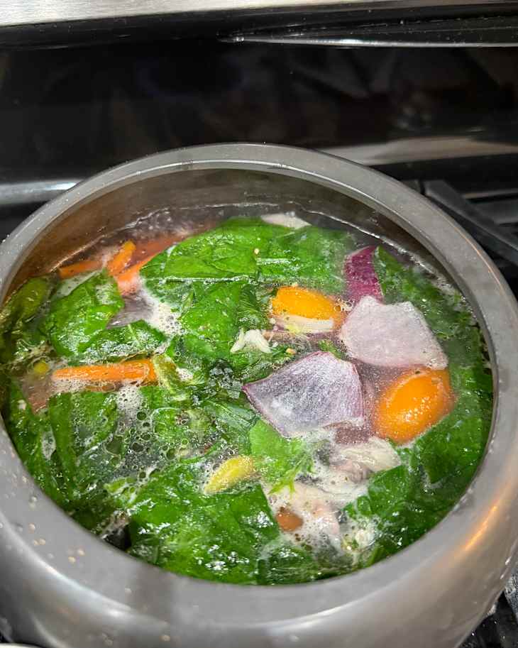 A pot of boiling soup with spinach, carrots, and purple onion on a stovetop.