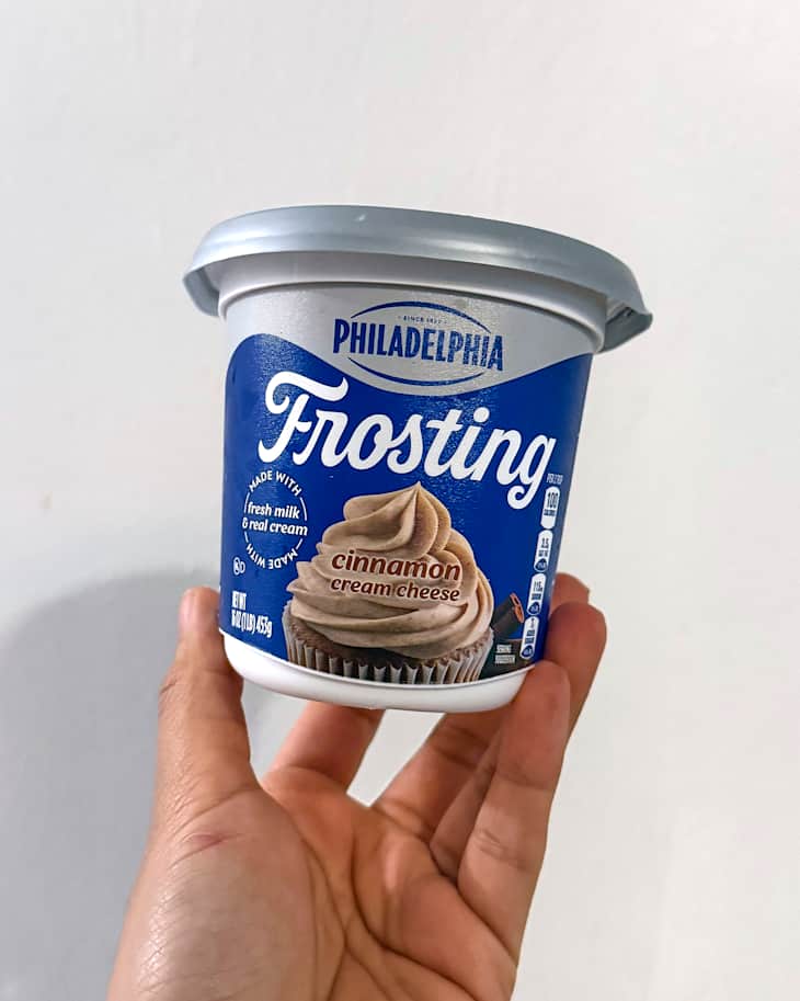 Cinnamon cream cheese frosting in a blue container held by a hand, with a cupcake image on the label.