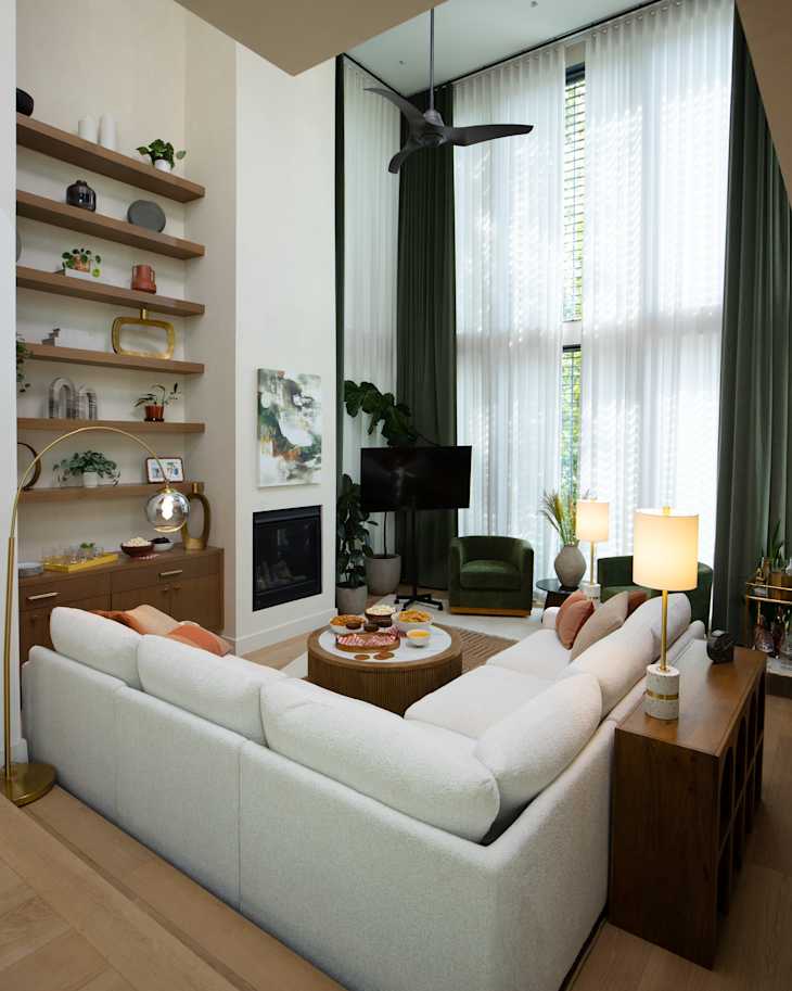 Cozy living room featuring a white sectional sofa, green armchairs, a wooden coffee table with snacks, and a modern fan.