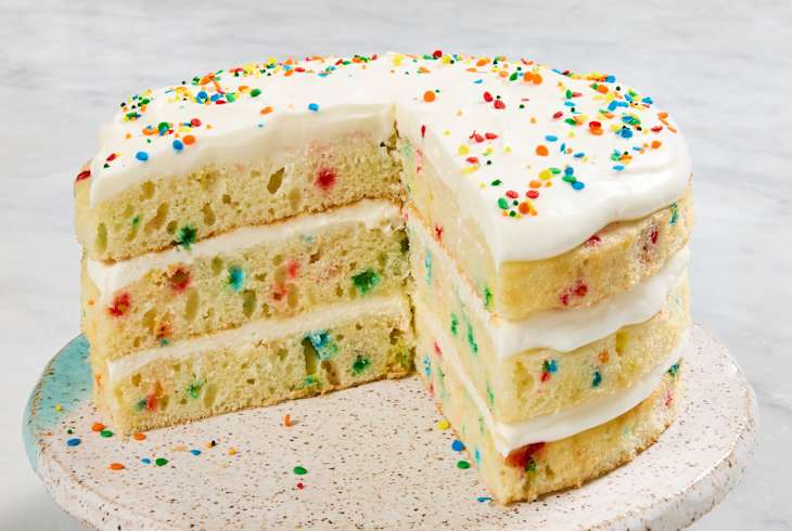 Layered cake with colorful sprinkles and white frosting on a speckled plate, partially sliced.