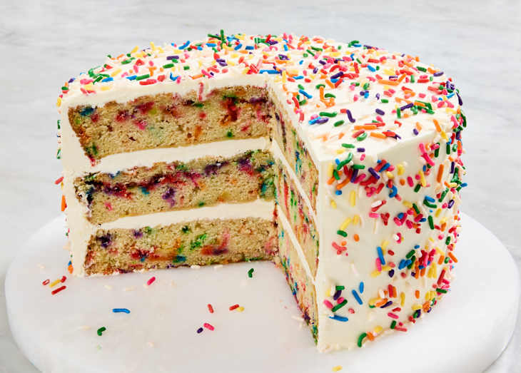 Layered cake with colorful sprinkles, creamy frosting, and a slice removed, displayed on a marble cake stand.