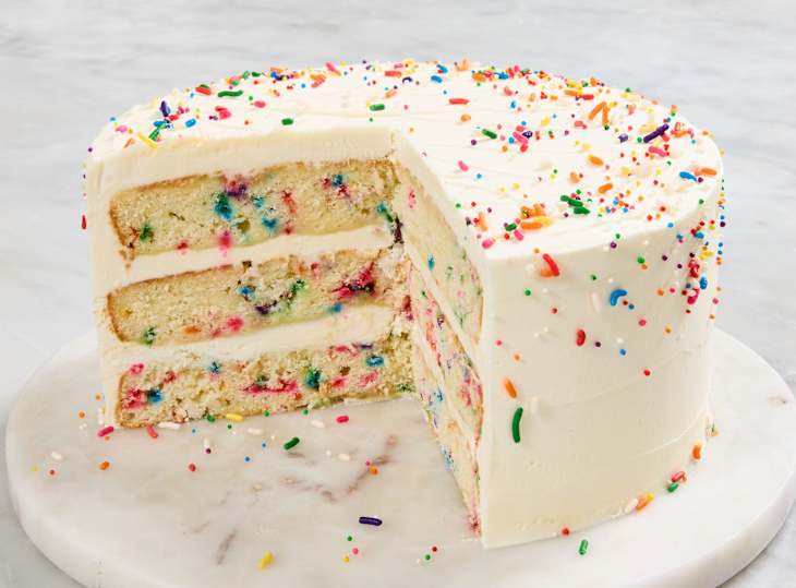 Layered vanilla cake with colorful sprinkles, frosted with white icing, on a marble cake stand.