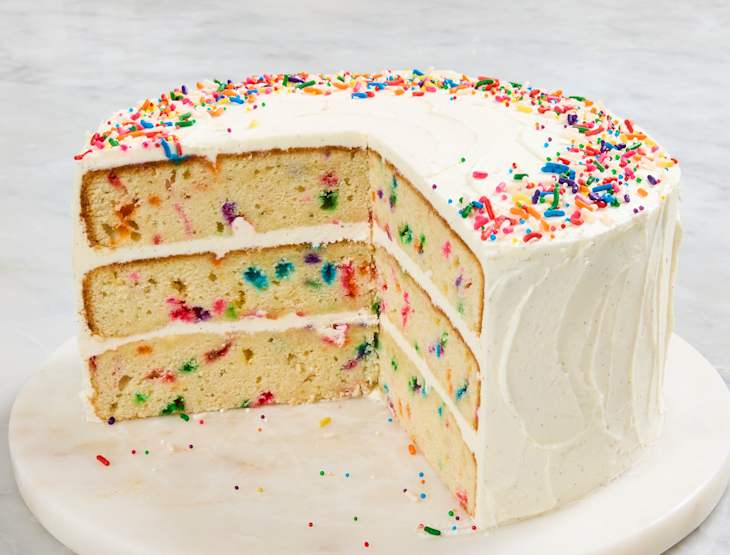 Layered vanilla cake with colorful sprinkles, frosted with white icing, displayed on a marble cake stand.
