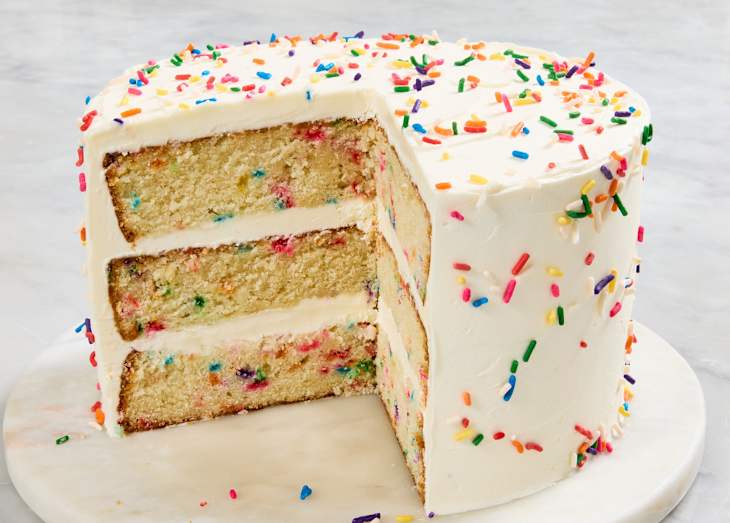 Layered vanilla cake with colorful sprinkles, frosted with white icing, partially sliced on a marble cake stand.