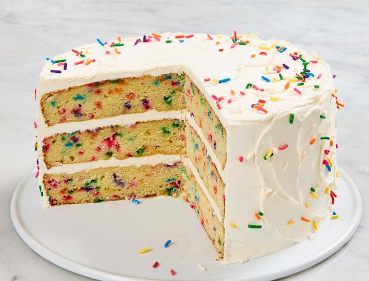 Layered vanilla cake with colorful sprinkles, frosted with white icing, partially sliced on a white plate.