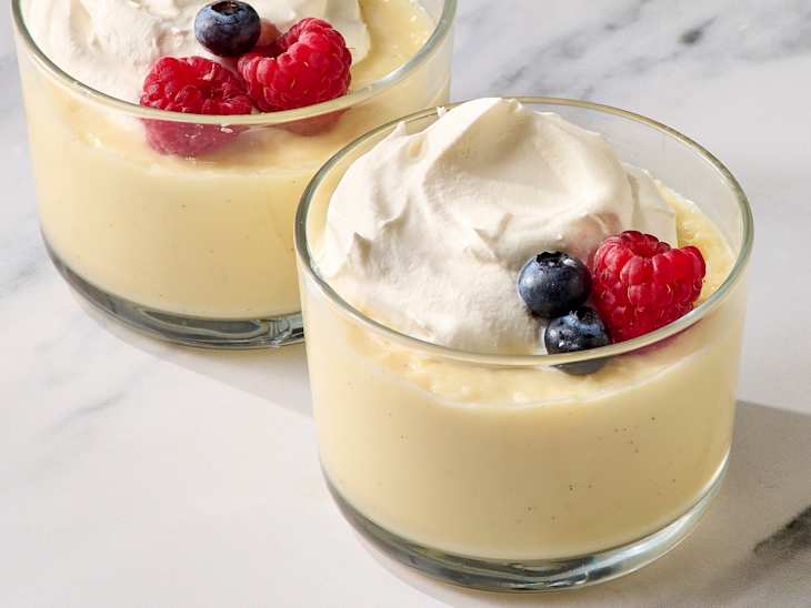 Two glass cups of creamy vanilla pudding topped with whipped cream, raspberries, and blueberries on a marble surface.