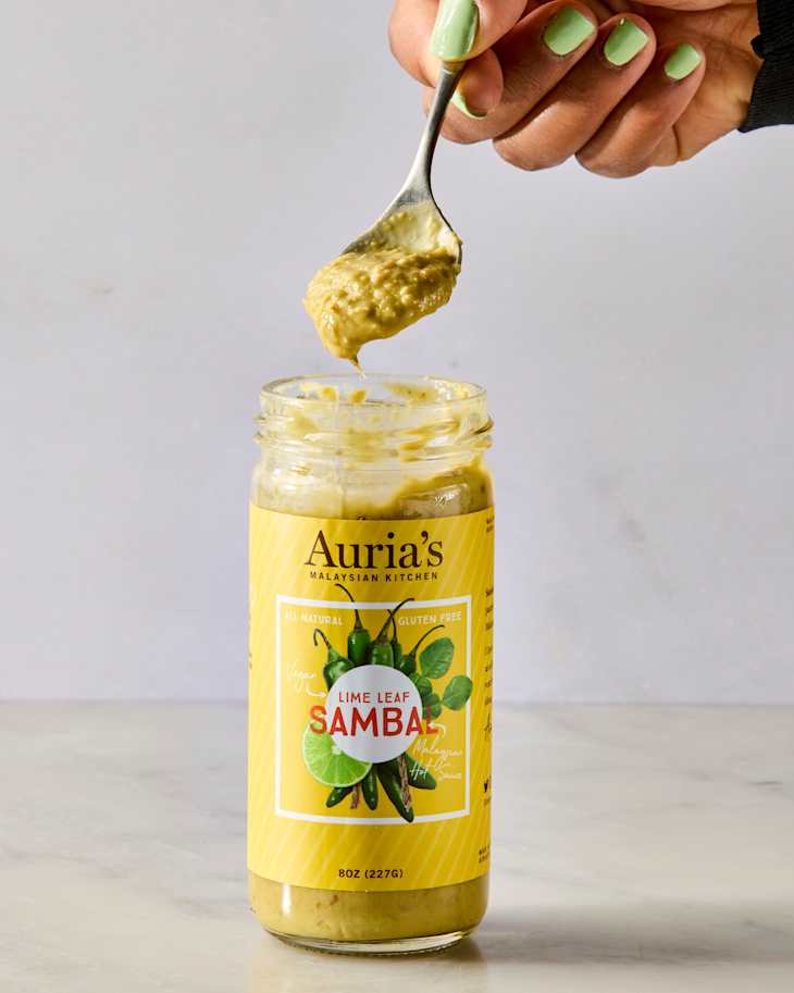 A hand holding a spoon with lime leaf sambal above a jar on a marble surface.