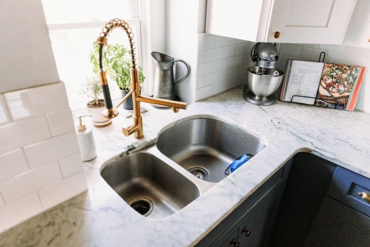 Marble kitchen countertop with double sink, brass faucet, potted herbs, stand mixer, and open cookbook.
