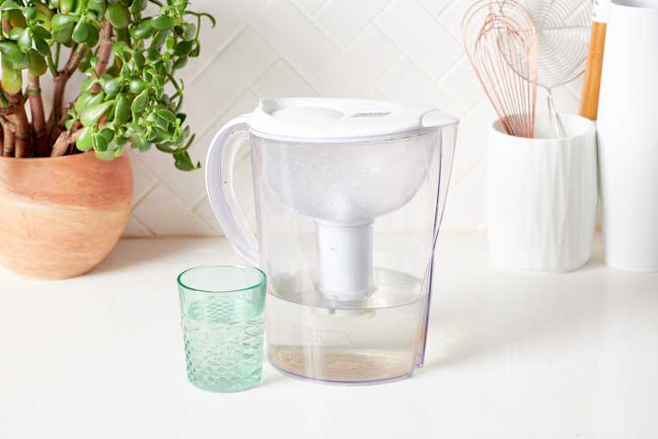 Water filter pitcher and green glass on a kitchen counter with a potted plant and utensils in the background.