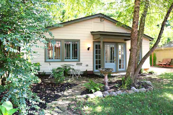 Charming cottage exterior with green trim, stone pathway, small table and chairs, and a birdbath surrounded by lush greenery.