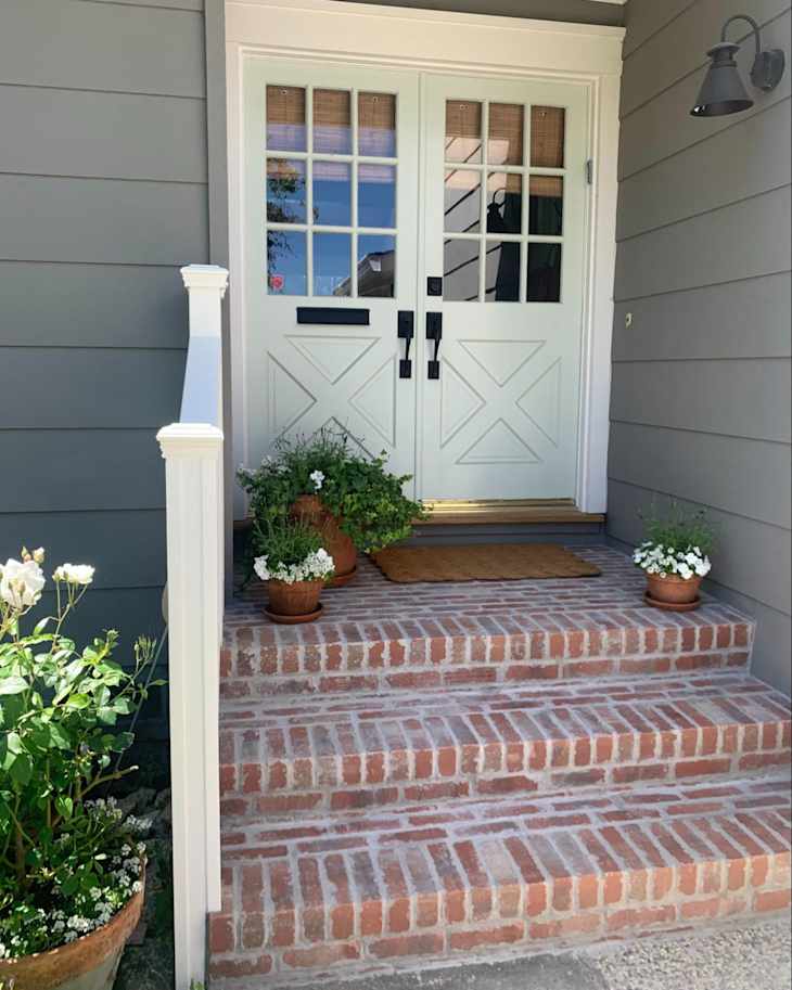 Front entrance with double doors, brick steps, potted plants, and a welcome mat.