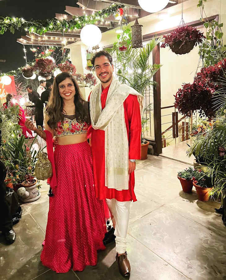 A couple dressed in traditional attire poses in a beautifully decorated outdoor space with plants and lights.