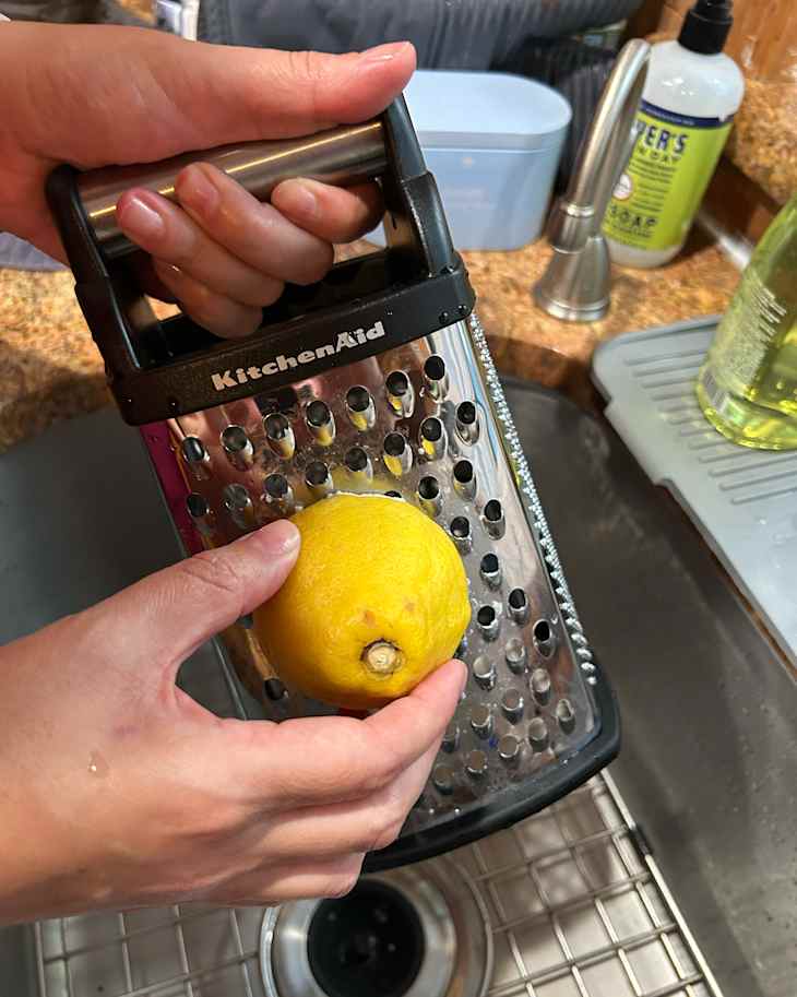 A person grating a lemon using a KitchenAid grater over a sink, with water and soap visible in the background.