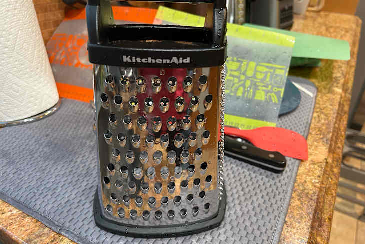 Stainless steel KitchenAid grater on a kitchen counter with a paper towel roll and colorful utensils nearby.