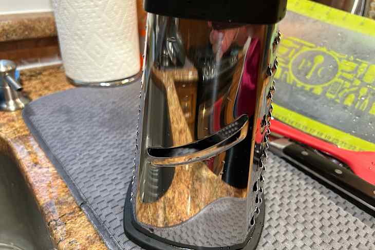 Stainless steel box grater on a gray mat, with a paper towel roll and kitchen utensils in the background.