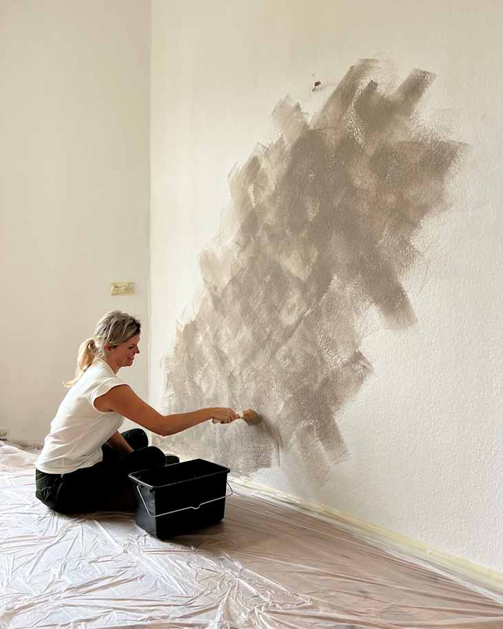 A woman painting a textured wall with a brush, sitting on a plastic-covered floor, with a paint bucket nearby.