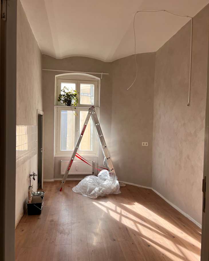 Room under renovation with a ladder, a potted plant by the window, and a pile of plastic sheeting on wooden flooring.