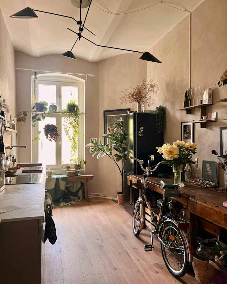 Cozy kitchen with wooden furniture, potted plants, a bicycle, and a large window letting in natural light.