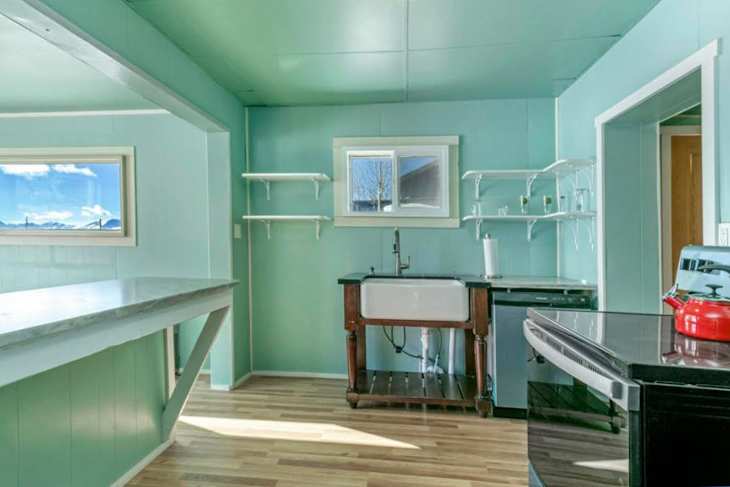 Bright turquoise kitchen featuring a farmhouse sink, wooden shelves, and modern appliances on a wood floor.