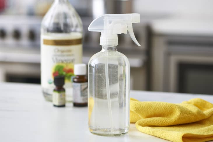 Spray bottle with cleaning solution, vinegar, essential oils, and yellow cloth on a kitchen counter.