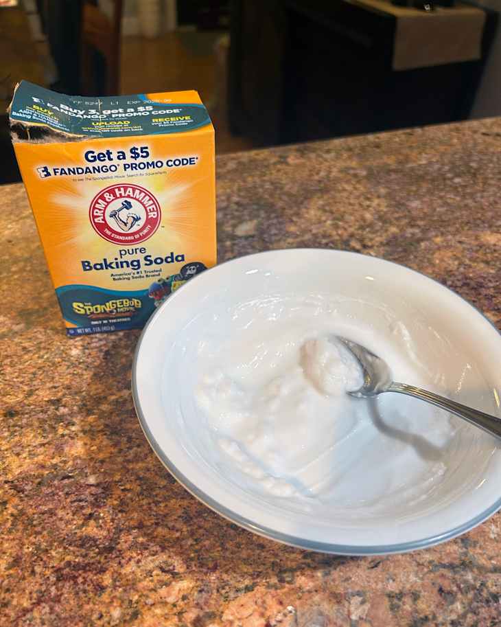 Baking soda box next to a white bowl containing a thick paste, on a speckled countertop.