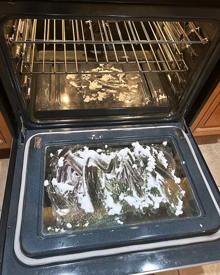 Oven interior with a glass door, racks, and a layer of baking soda paste on the bottom and door
