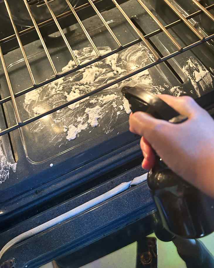 A hand holding a spray bottle of vinegar cleaning the interior of an oven with baking soda paste on the bottom and oven racks.