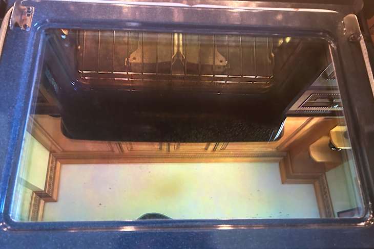View of a clean oven door with a glass panel reflecting kitchen cabinetry and a wire rack inside.
