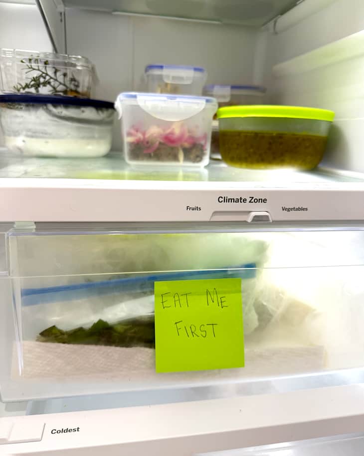Refrigerator shelf with various food containers, including a note saying "EAT ME FIRST" on a bag of greens.