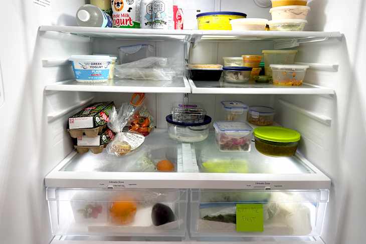 Refrigerator interior filled with various food containers, fruits, vegetables, and a note saying "EAT ME FIRST."