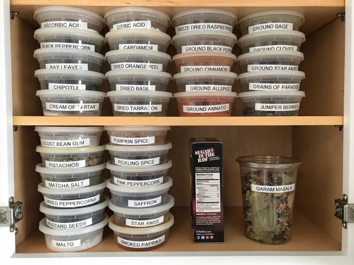 Kitchen cabinet with labeled spice containers, including cardamom, juniper berries, and garam masala.