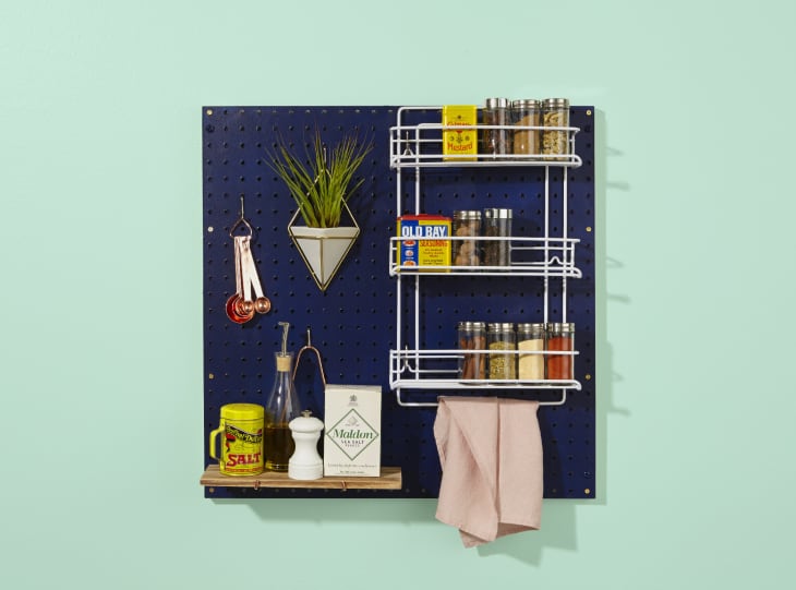 Navy pegboard with spice rack, measuring spoons, plant, and shelf holding salt, oil, and pepper grinder.