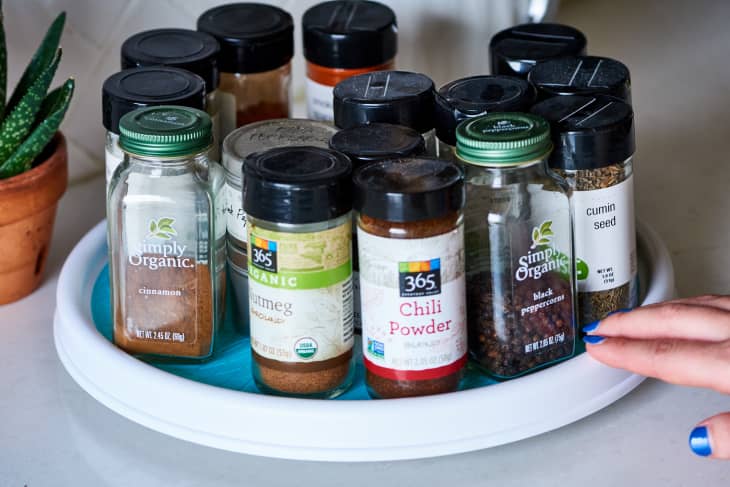 Assorted spice jars on a white lazy Susan, including cinnamon, chili powder, and cumin seed, next to a small potted plant.