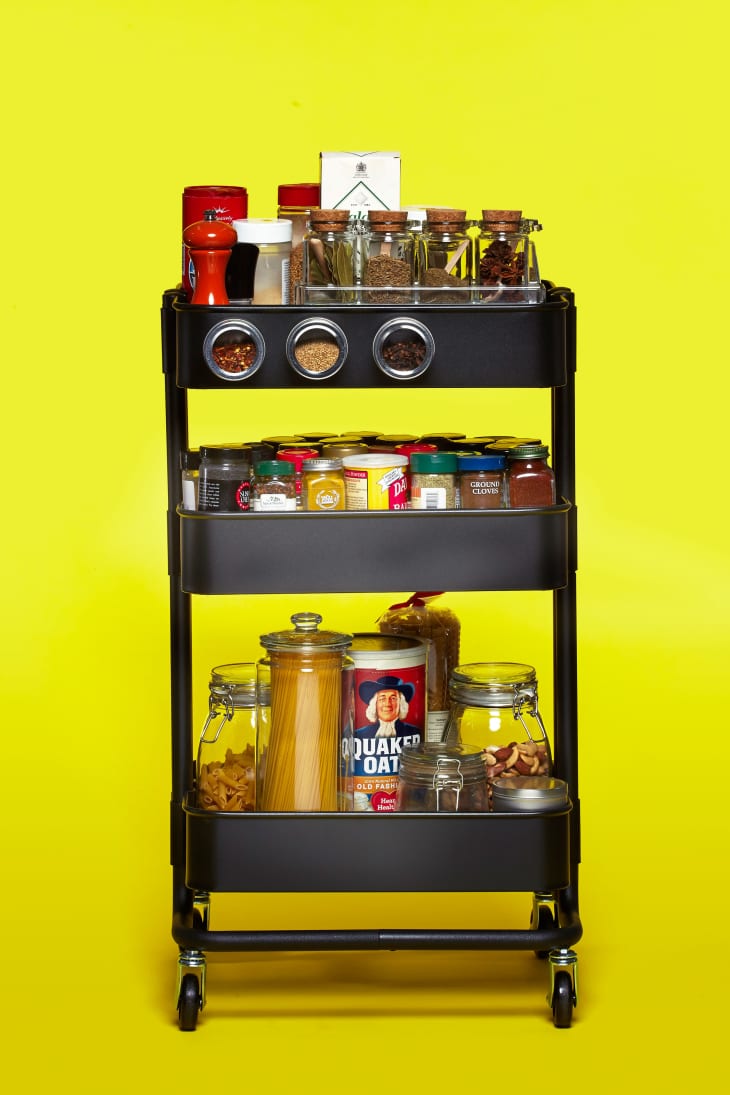 Black kitchen cart with spices, pasta, oats, and jars on a bright yellow background.
