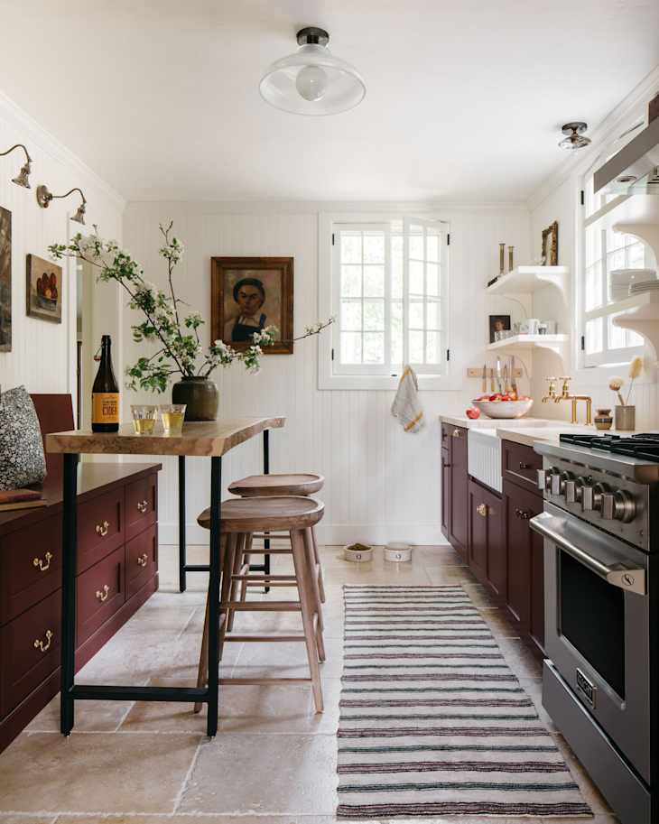Cozy kitchen featuring burgundy cabinets, a wooden bar table, two stools, and a striped rug, with floral decor and artwork.