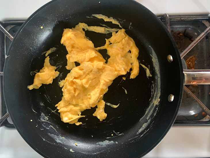 Scrambled eggs in an All-Clad skillet on a stovetop, with some egg residue on the surface.