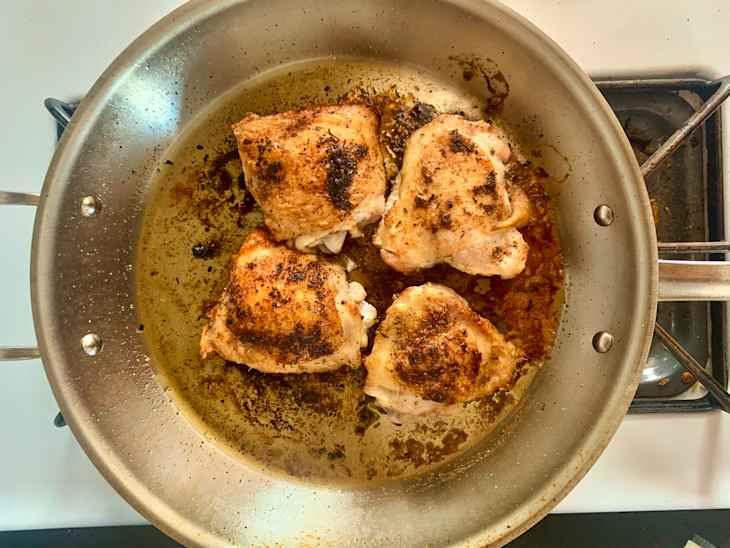 Four browned chicken thighs cooking in an All-Clad skillet with oil and seasoning.
