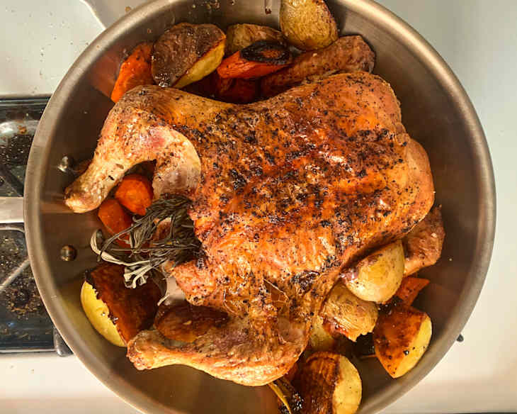 Roasted chicken with crispy skin surrounded by assorted vegetables, including potatoes and carrots in an All-Clad skillet