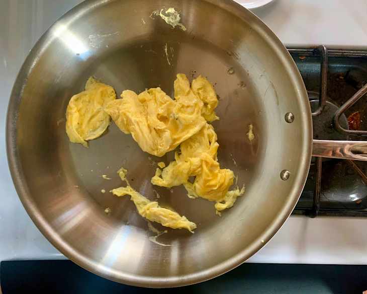 Scrambled eggs in an All-Clad skillet on a stovetop, with some pieces slightly browned.