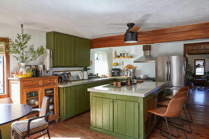 Modern kitchen with green cabinets, a marble island, wooden accents, and a dining table with a patterned chair.