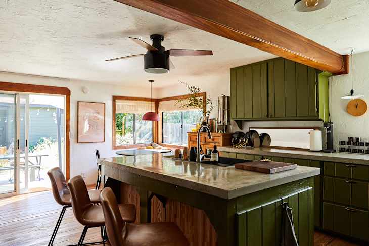 Modern kitchen with green cabinets, concrete countertops, wooden beams, and a dining area visible through sliding doors.