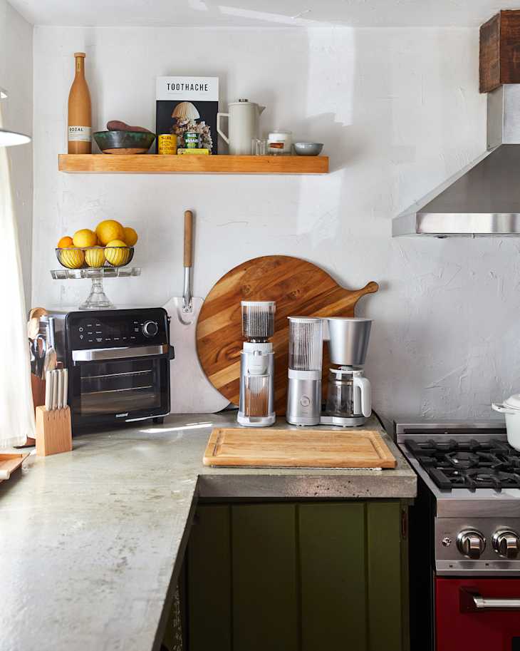 Modern kitchen corner with a concrete countertop, coffee maker, and an oven, featuring a wooden cutting board and decorative shelf.