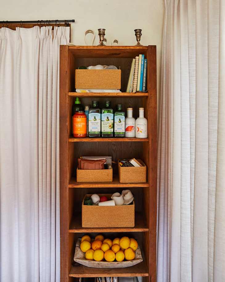 Wooden shelf with various bottles, woven baskets, and a bowl of lemons, next to white curtains.