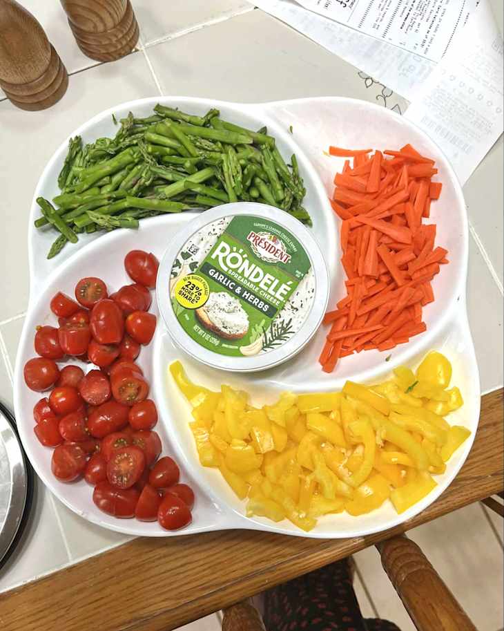 Chopped vegetables including asparagus, cherry tomatoes, carrots, and yellow bell peppers arranged on a white platter with garlic herb cheese.