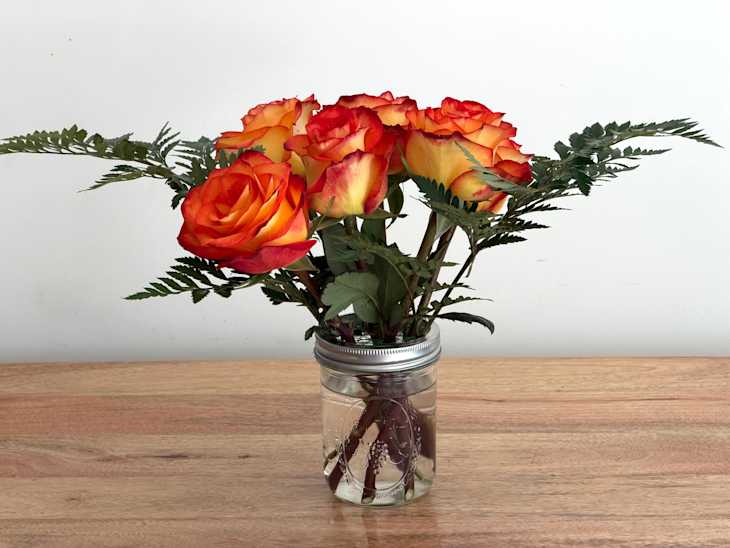 Vibrant orange and yellow roses arranged in a mason jar with green ferns on a wooden surface.