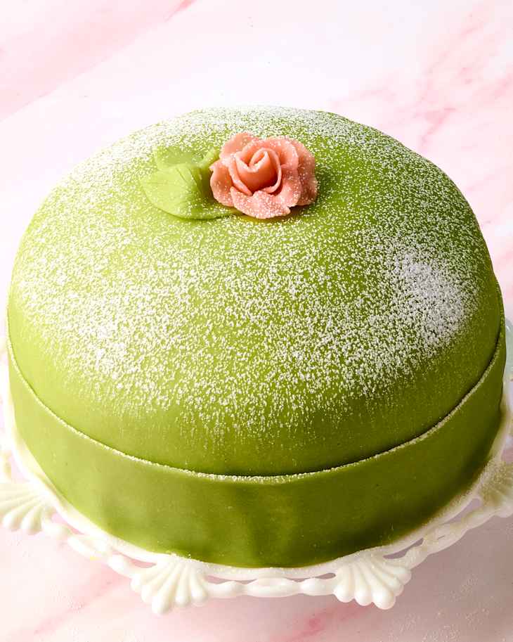 Green cake with a smooth finish, topped with a pink rose and leaf, dusted with powdered sugar on a decorative plate.